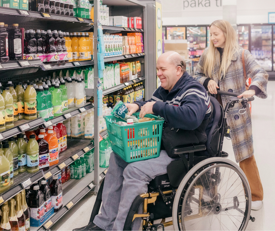 Phil, sits in his wheelchair at a grocery store. His Independent Living Skills Specialist is smiling behind him, holding the wheelchair handles. Phil, sits in his wheelchair at a grocery store. His Independent Living Skills Specialist is smiling behind him, holding the wheelchair handles.