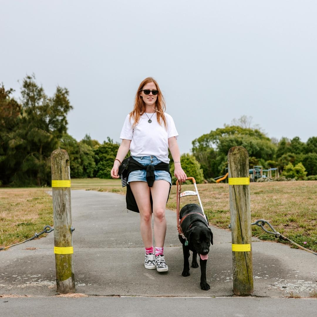 A woman in a white t-shirt, shorts, and sunglasses walks forward with a black guide dog on a paved path. Trees and a playground are in the background.