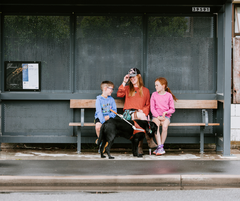 Laura sitting at a bus stop with her two young children, and her black guide dog Ryder Laura sitting at a bus stop with her two young children, and her black guide dog Ryder