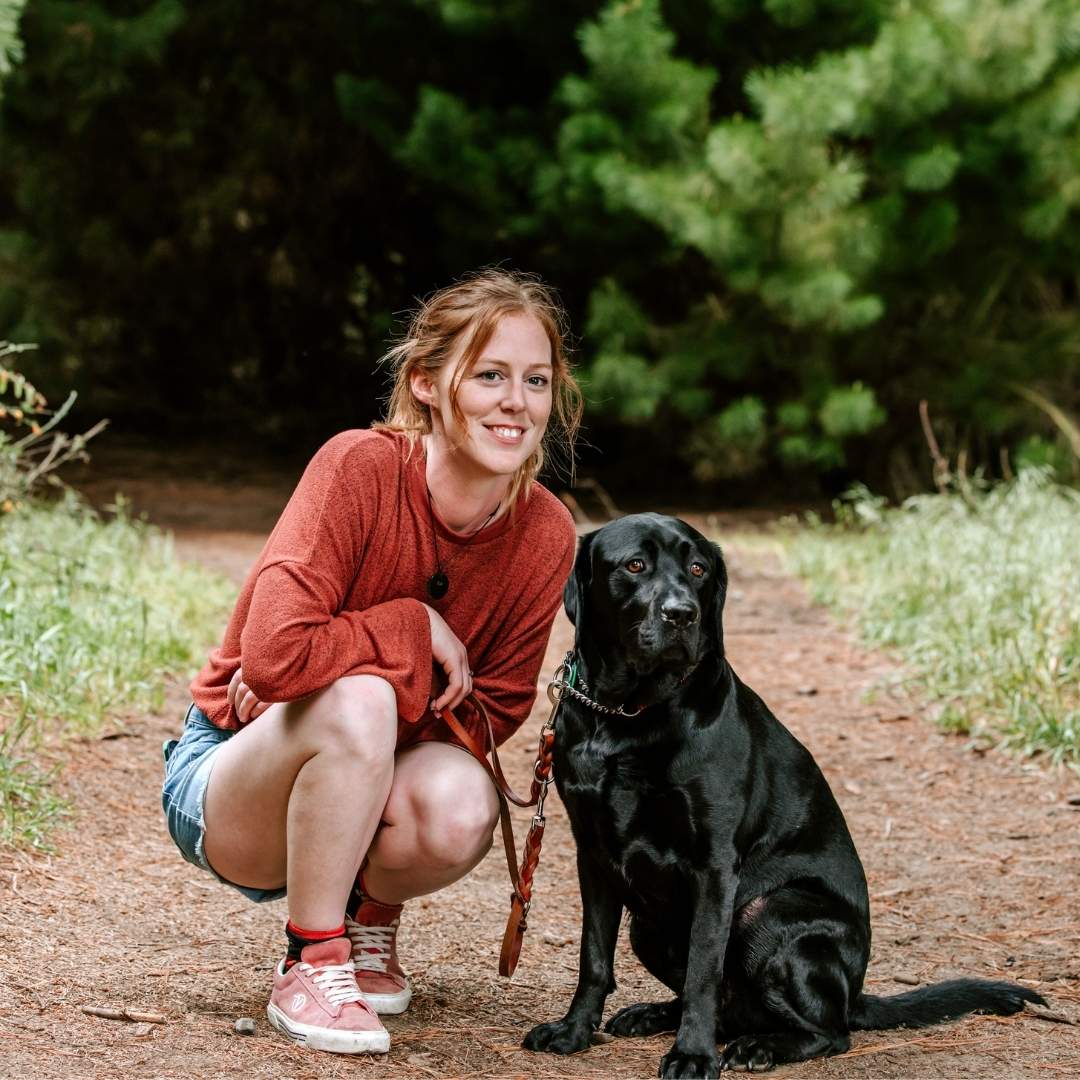 A woman and a black Lab sit on a dirt path, smiling at the camera. Greenery blurs in the background.