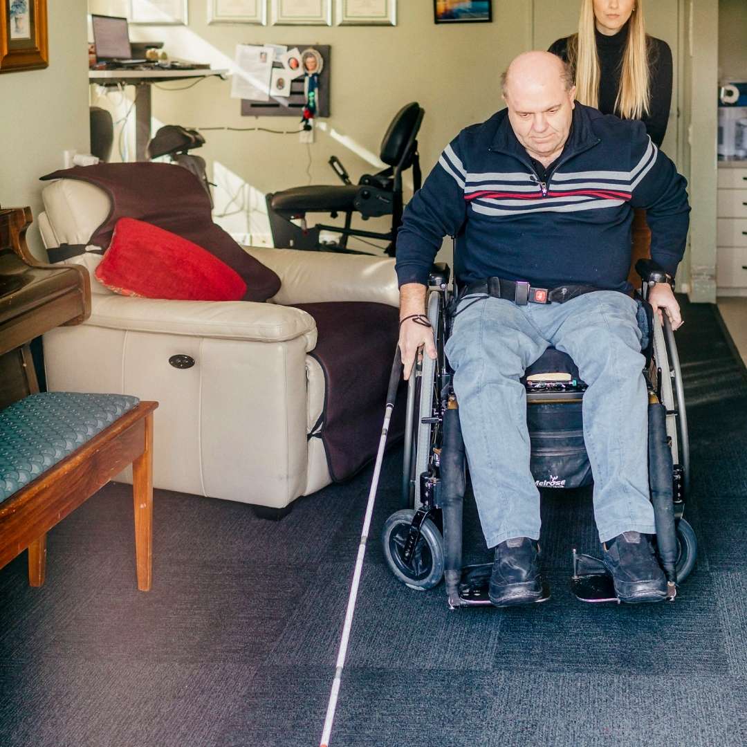 A man in a wheelchair being pushed by a lady, in his home. The man also has a white cane A man in a wheelchair being pushed by a lady, in his home. The man also has a white cane