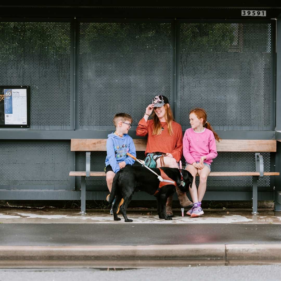 A woman and two children sit on a bench at a bus stop, with a black guide dog at their feet. The woman adjusts her cap, looking at the boy. The bus stop has a grey metal back panel and a route map.